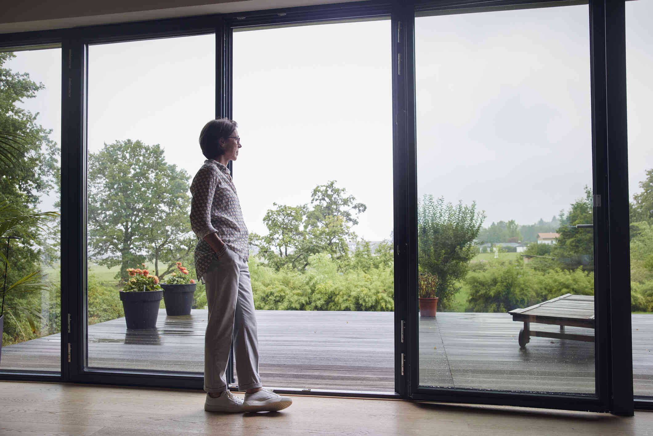 A middle aged woman stands in her home near an open glass door and gazes out into her backyard with a sad expression.